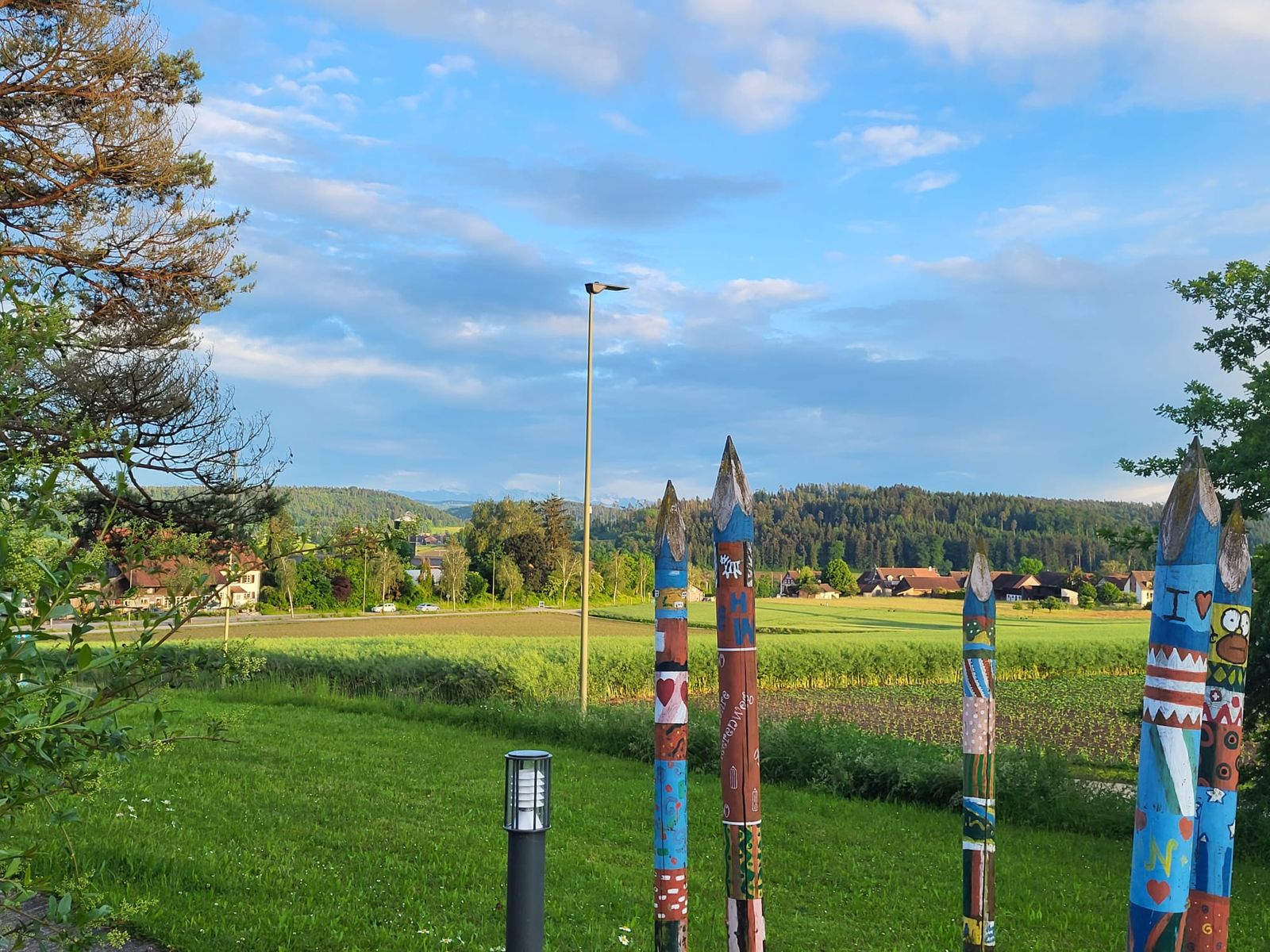 Bemalte Holzpfosten am Rand des Schulareals Heerenweg, dahinter Felder und Aesch